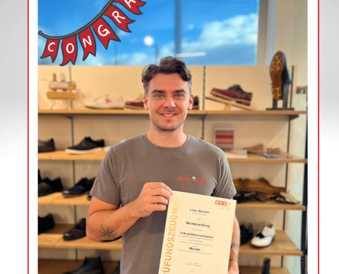 Lukas steht lächelnd in unserer Filiale und hält stolz sein Meisterprüfungszeugnis zum Orthopädieschuhmacher in den Händen. Über ihm hängt eine rote Girlande mit dem Schriftzug „Congrats“. Im Hintergrund sind Regale mit Schuhen zu sehen.