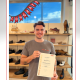 Lukas steht lächelnd in unserer Filiale und hält stolz sein Meisterprüfungszeugnis zum Orthopädieschuhmacher in den Händen. Über ihm hängt eine rote Girlande mit dem Schriftzug „Congrats“. Im Hintergrund sind Regale mit Schuhen zu sehen.
