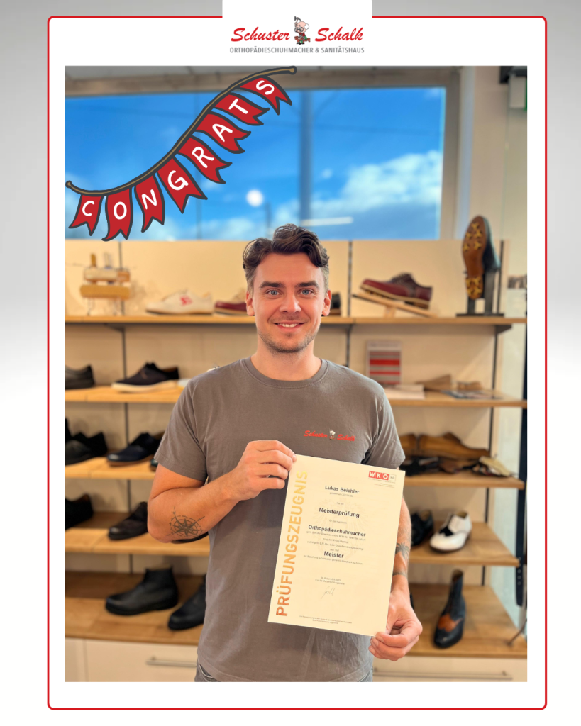 Lukas steht lächelnd in unserer Filiale und hält stolz sein Meisterprüfungszeugnis zum Orthopädieschuhmacher in den Händen. Über ihm hängt eine rote Girlande mit dem Schriftzug „Congrats“. Im Hintergrund sind Regale mit Schuhen zu sehen.